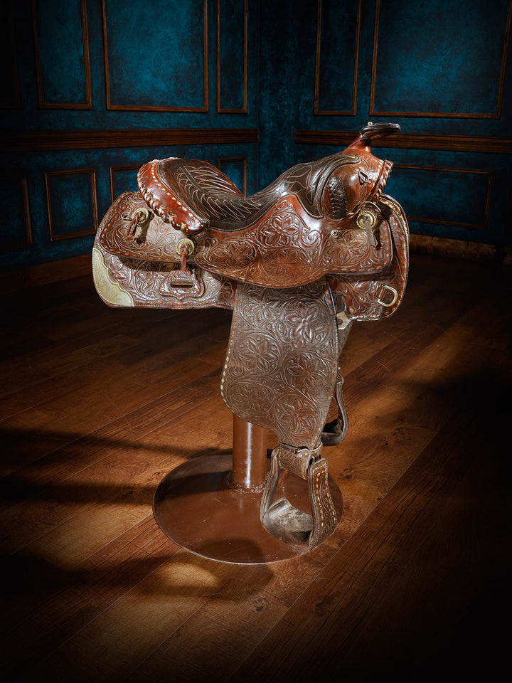 A unique bar stool designed with a richly tooled leather Western saddle, mounted on a sturdy metal base, offering a rustic and authentic cowboy aesthetic.