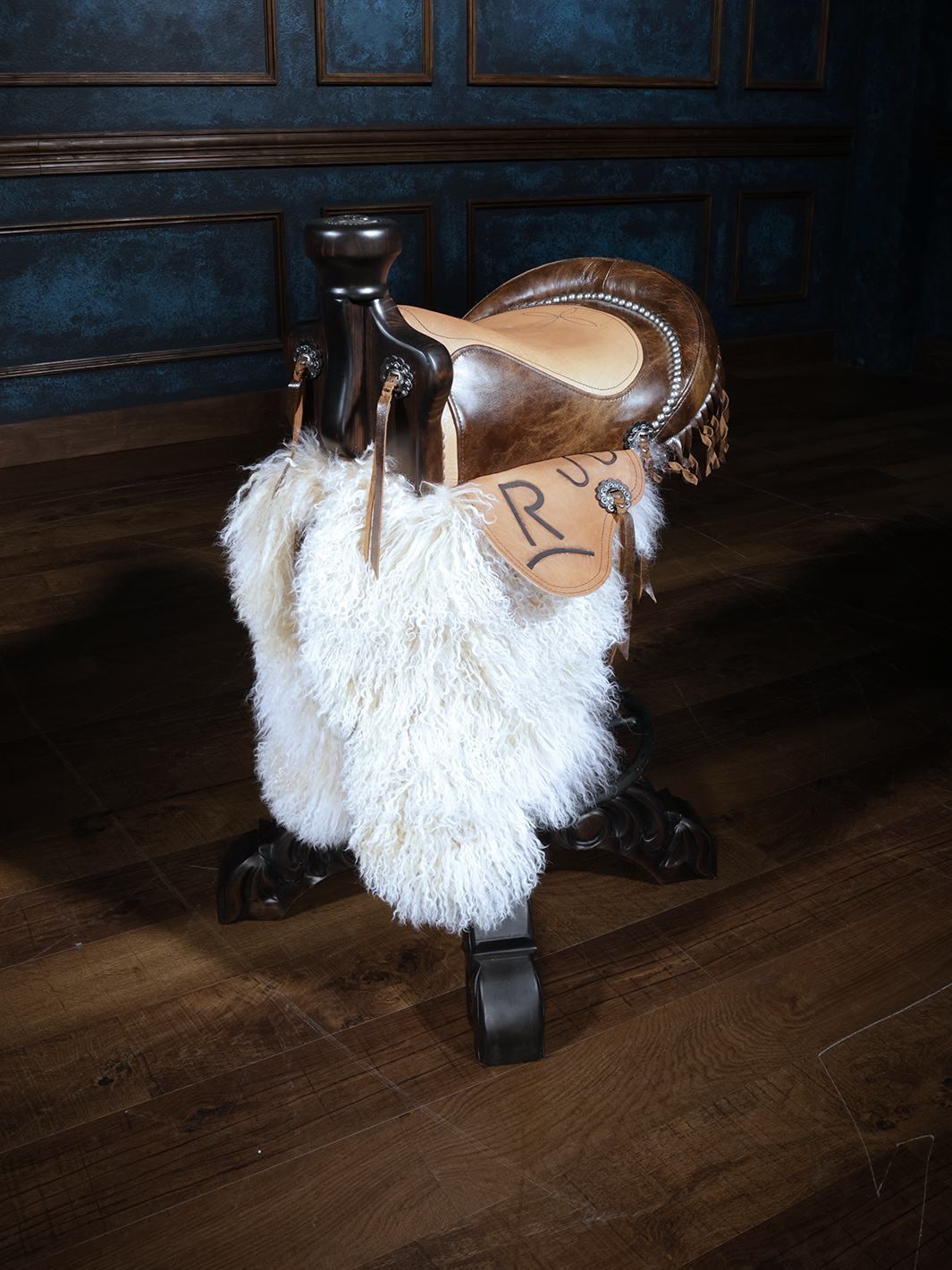 A Western style saddle stool with a genuine leather seat and white Tibetan wooly chaps, displayed in a room with wooden flooring and blue walls.