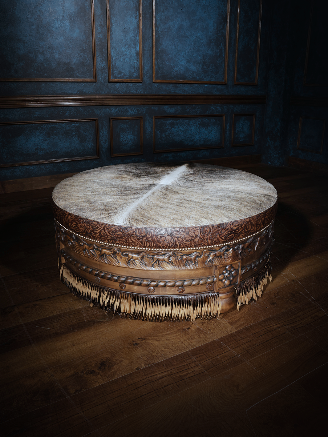 A brown leather ottoman with fringe detailing, placed on a wooden floor with a patterned blue wall in the background.