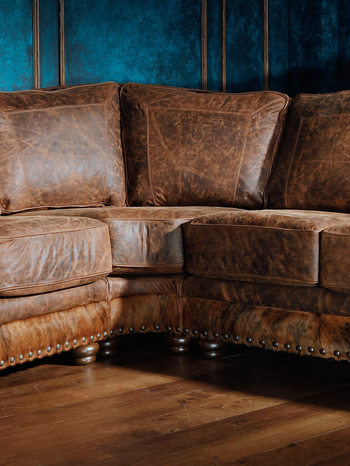 Close-up of the corner section of a western-style sectional sofa, featuring distressed brown leather upholstery, hair-on-hide accents, and nailhead trim.